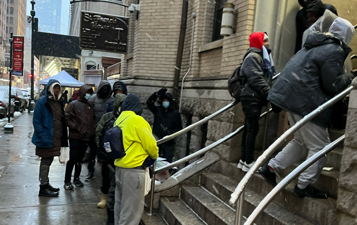 Several people in winter clothing wait along a sidewalk and on stairs to a building on a city street on a wet and snowy day.