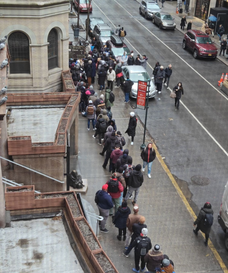 Dozens of people in winter clothing wait along a wet city sidewalk to get into a building.