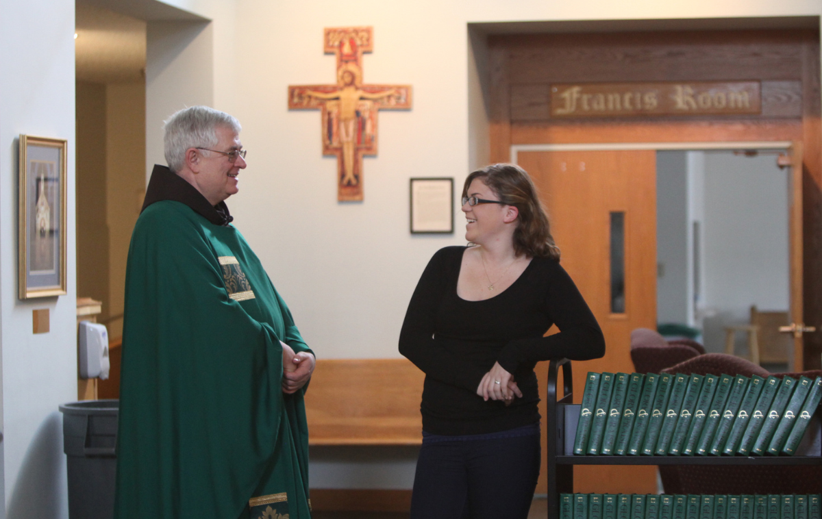 A man in vestments and glasses talks with a student who is also wearing glasses.