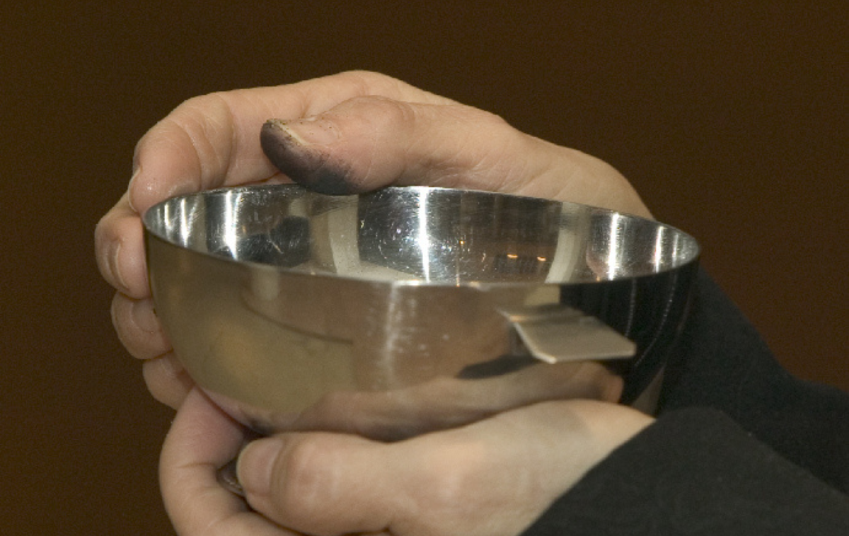 A man holds a bowl of ashes. His thumb is black with ash.