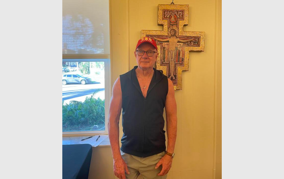 A 78-year-old friar stands in front of a San Damiano Cross hanging on a wall.