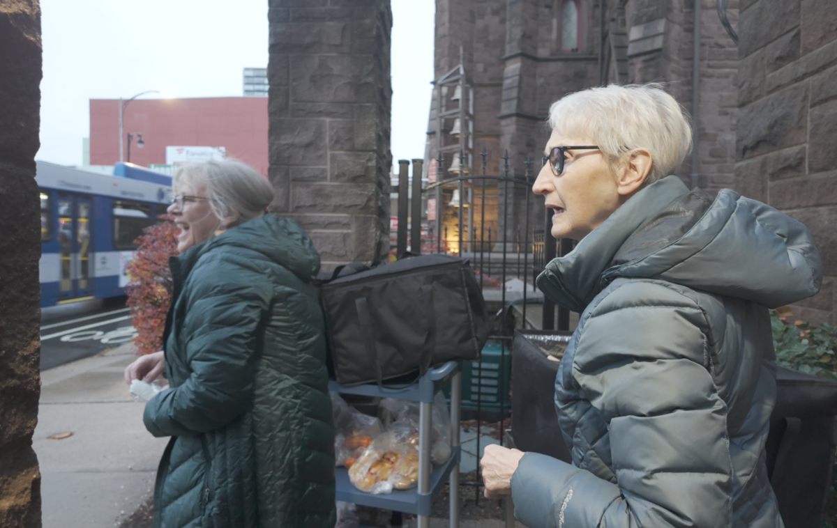 Two women in winter coats laugh as they push a cart loaded with food.