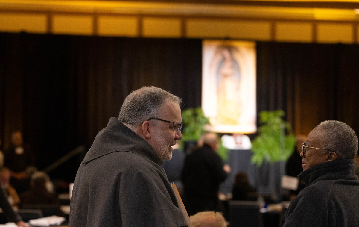 Four men stand in a room beside a plant and in front of a crucifix. There is a statue of Jesus Christ behind them.