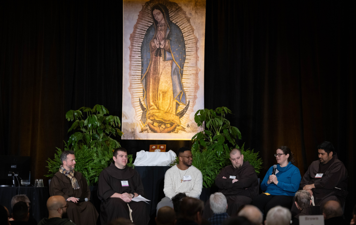 A group of five young men and a young woman sit on a stage. A woman and one of the men are wearing street clothes – the rest are wearing Franciscan habits.