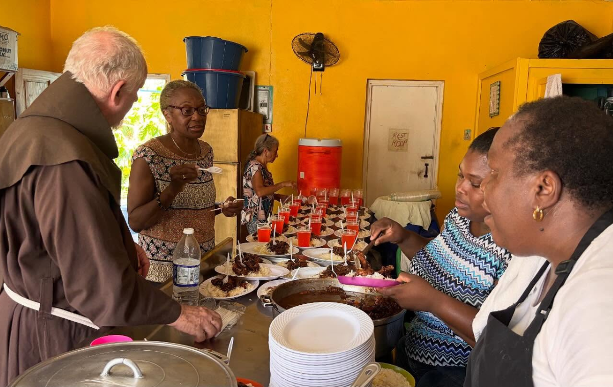 A friar welcomes three people who are picking up bags of food.