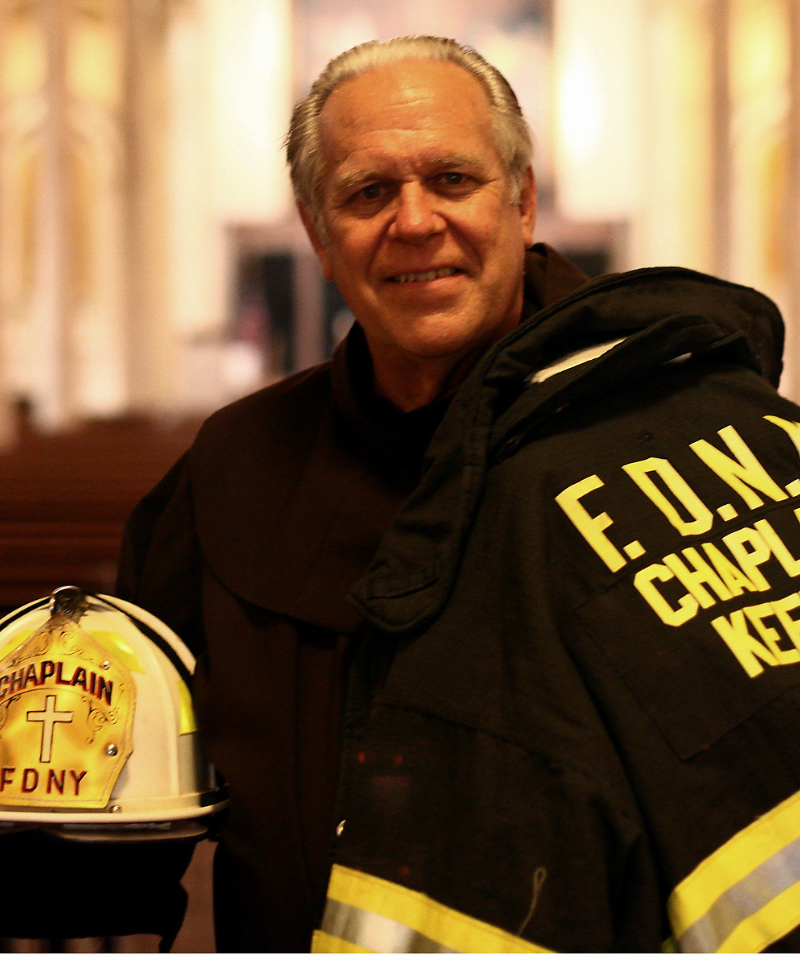 A smiling man holds a firefighter jacket and helmet in a church.