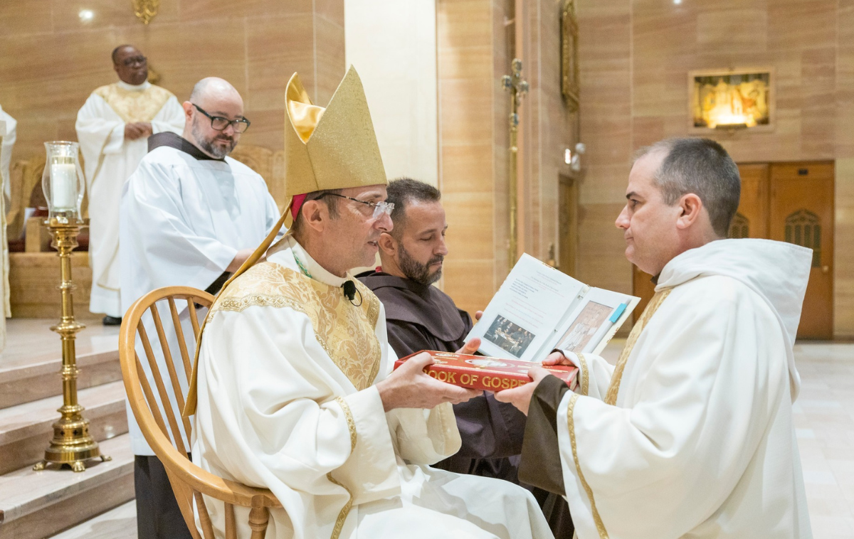 A seated man in priest vestments holds a book with another man in vestments. A bearded man in a friar habit sits next to him. Two more men in vestments stand behind the group.