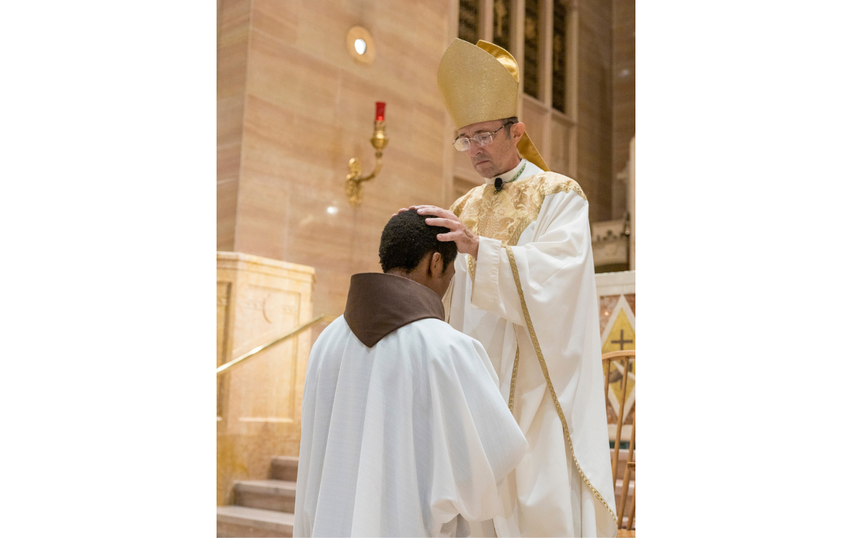 A man in a friar habit and vestments kneels before another man in vestments and a cap who is laying hands on the kneeling man's head.