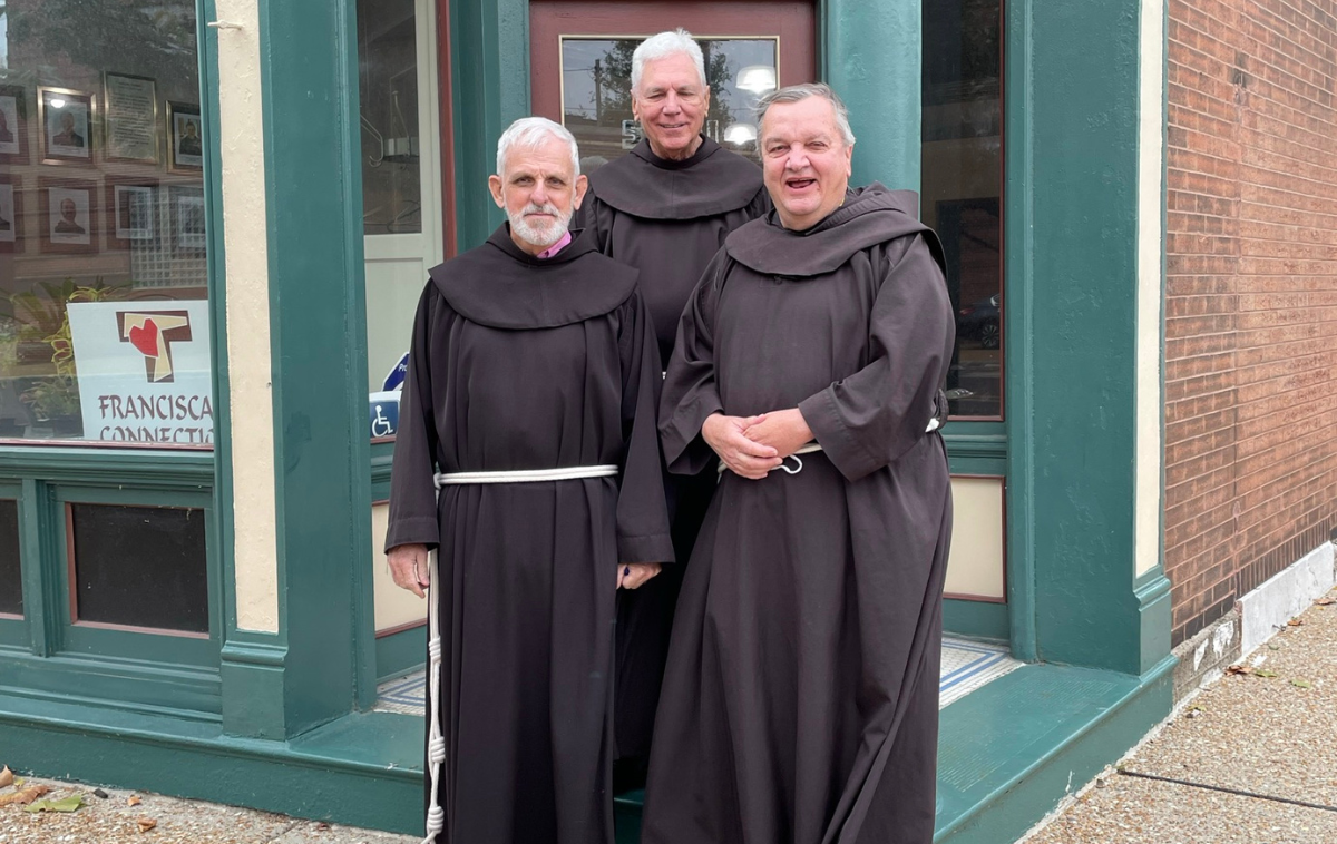 Three friars stand in front of a small storefront