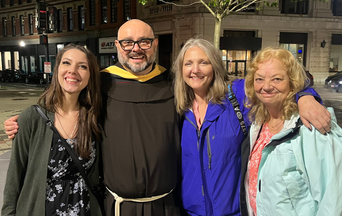 A smiling man with eyeglasses, wearing a friar habit and yellow sash, poses for an outdoor photo with three women.