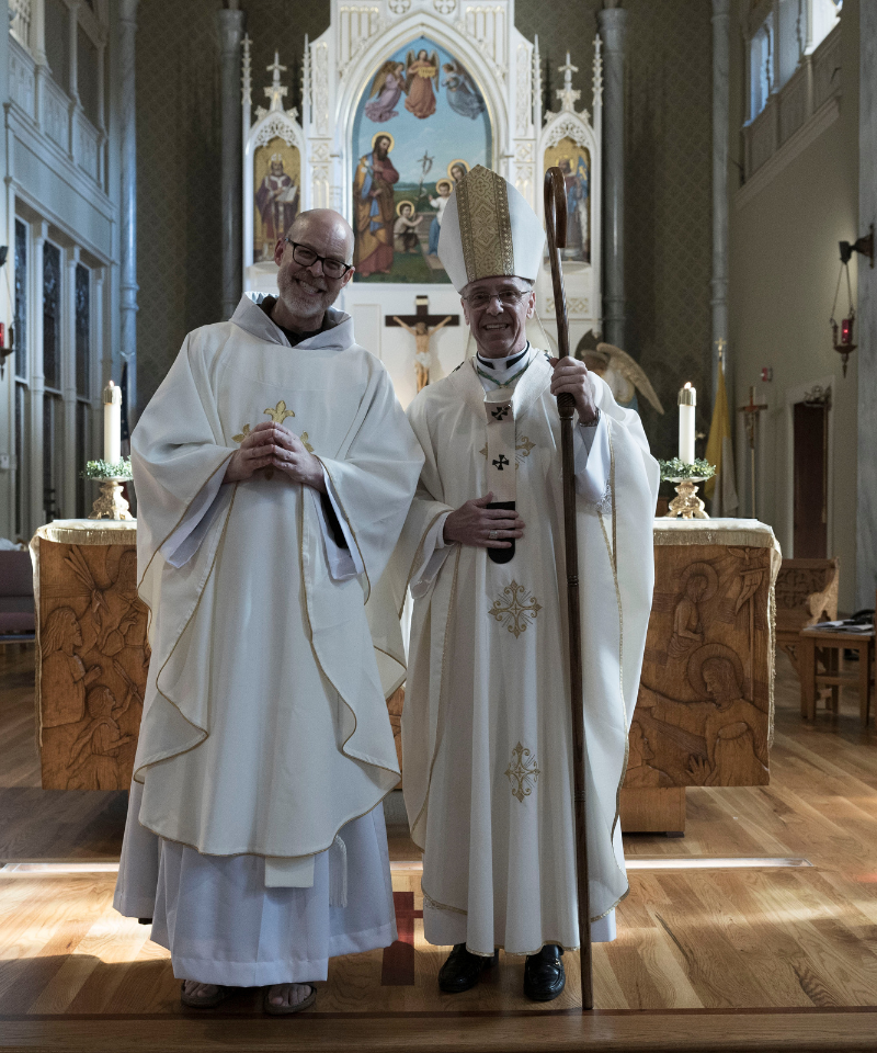 A priest stands with an archbishop in the front of a Catholic Church.