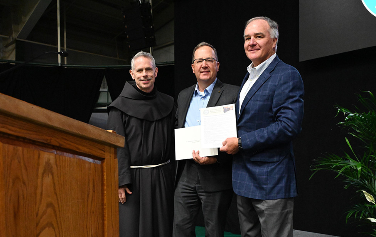 One man in a friar habit and two men in sports jackets pose with documents in front of a podium.