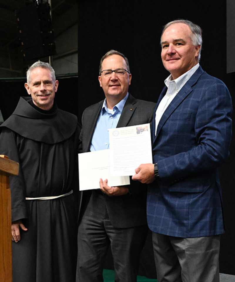 A man in a friar habit and two men in sports jackets pose with documents in front of a podium.