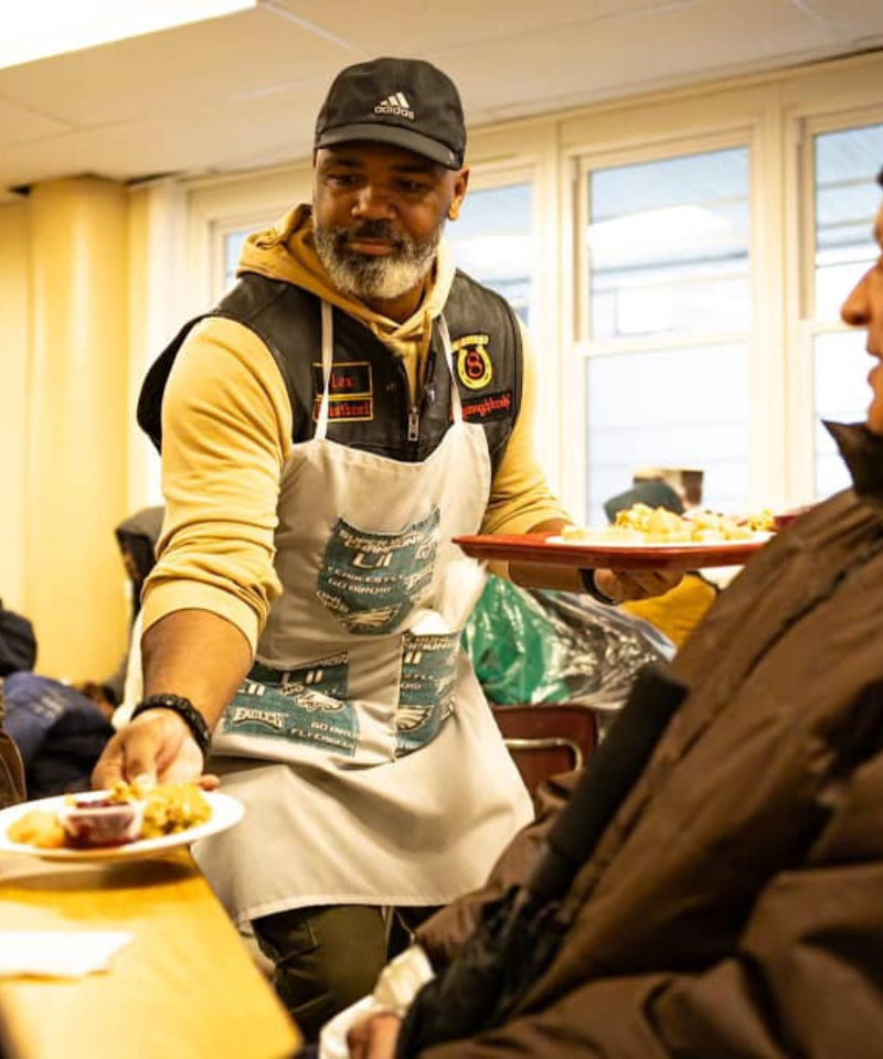 A bearded man wearing a baseball cap and apron serves food on plates to a man seated at the table. The man seated is wearing a brown coat.