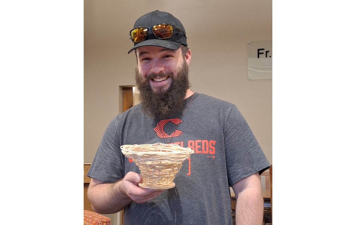 A bearded, smiling man wearing a baseball cap with sunglasses on it and a Cincinnati Reds t-shirt holds a weaved basket.