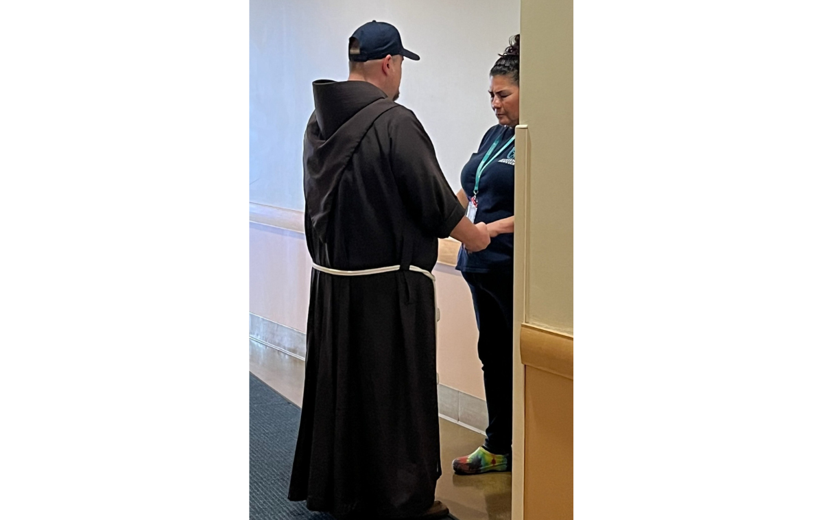 A man wearing a friar habit and a baseball cap holds hands with a woman in a blue t-shirt.