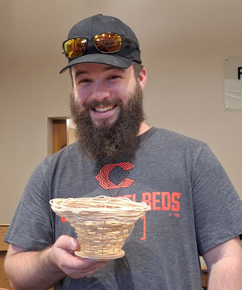 A bearded, smiling man wearing a baseball cap with sunglasses on it and a Cincinnati Reds t-shirt holds a weaved basket.