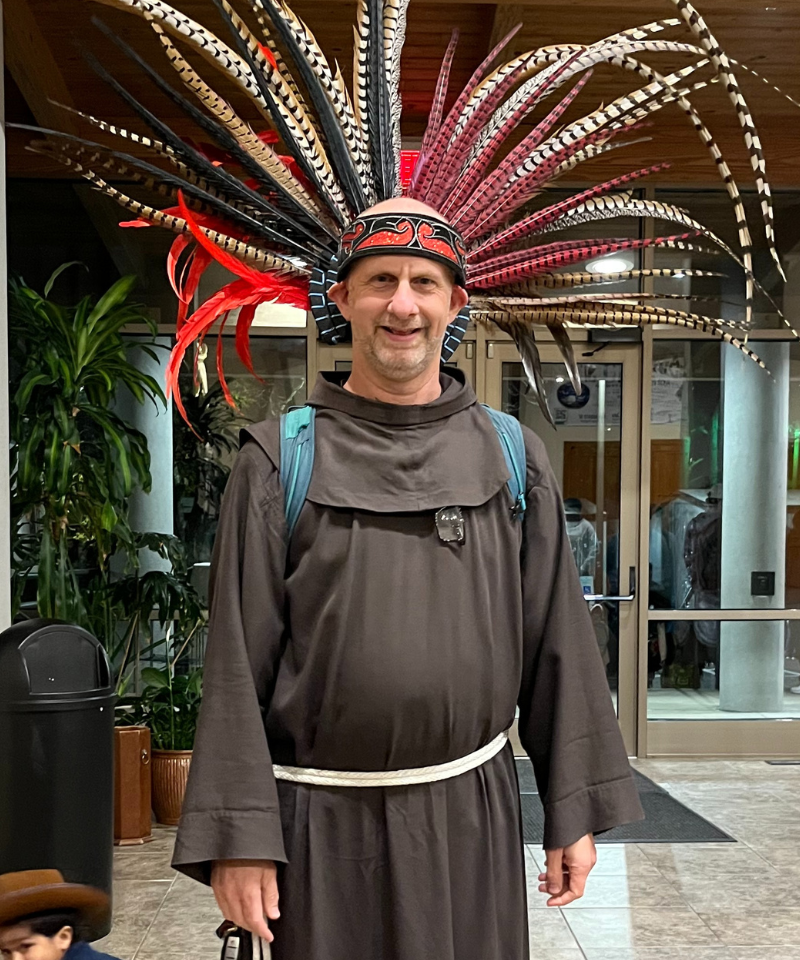 A man wearing a friar habit wears a headdress of red and yellow feathers.