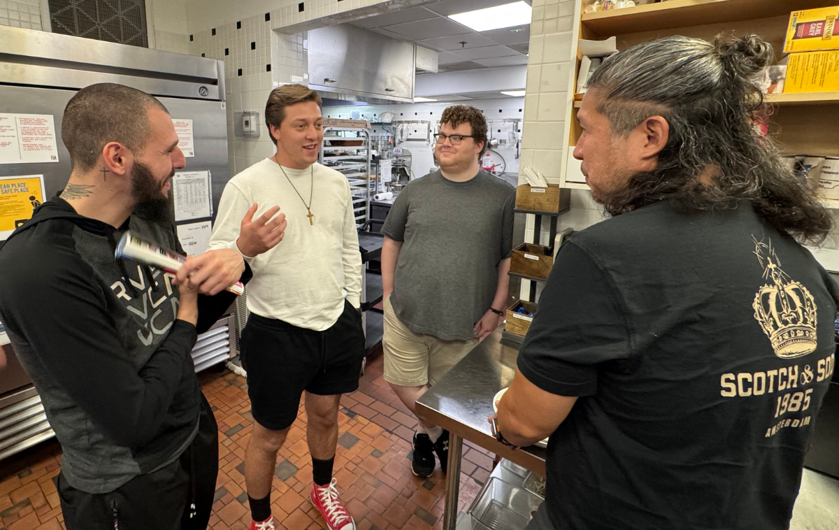 Four men stand while having a discussion in a kitchen. There are refrigerators and food carts in the background.