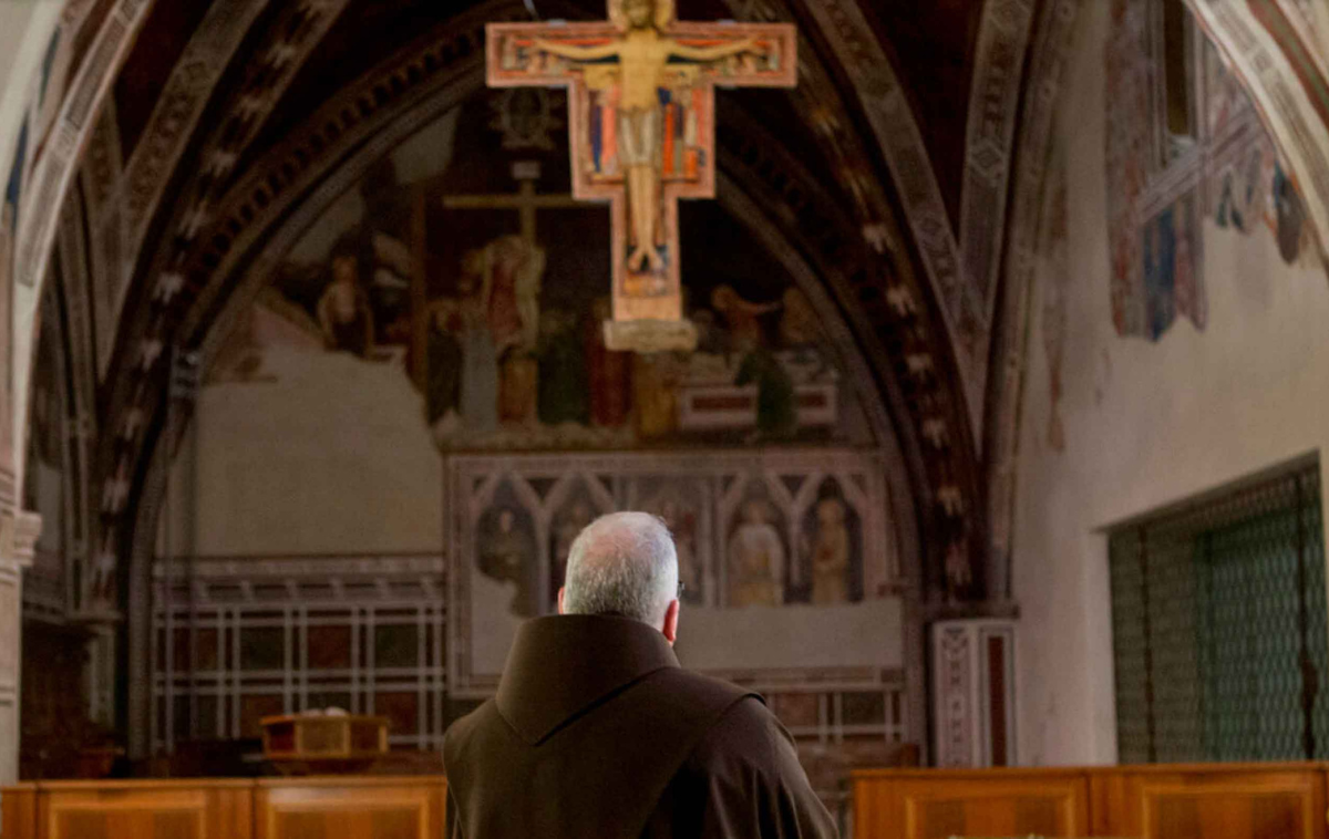 A friar stands before the San Damiano cross.